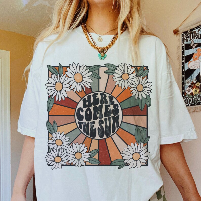 May include: White t-shirt featuring a retro graphic design with a sunburst and the text "Here Comes The Sun" in a groovy font. The graphic is surrounded by colourful flowers.
