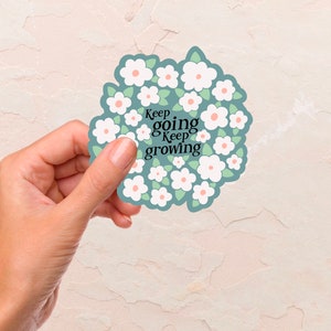 May include: A white sticker with a circle of white flowers with pink centers and green leaves. The text "Keep going Keep growing" is in the center of the sticker.