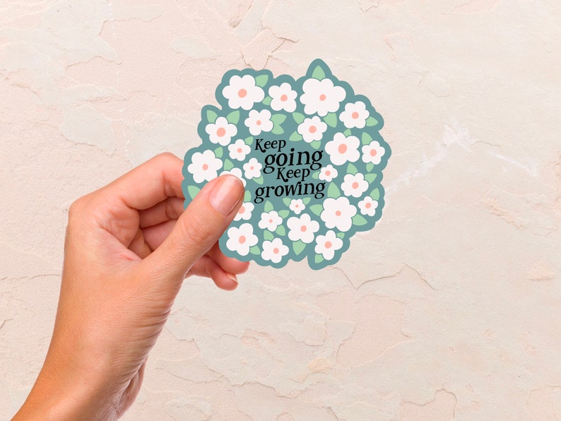 May include: A white sticker with a circle of white flowers with pink centers and green leaves. The text "Keep going Keep growing" is in the center of the sticker.