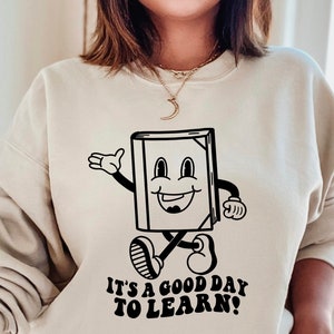 May include: A beige sweatshirt with a black and white illustration of a smiling book character with the text "It's a good day to learn!"