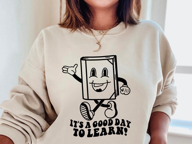 May include: A beige sweatshirt with a black and white illustration of a smiling book character with the text "It's a good day to learn!"