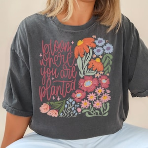 May include: A dark gray t-shirt with a floral design and the text "bloom where you are planted".