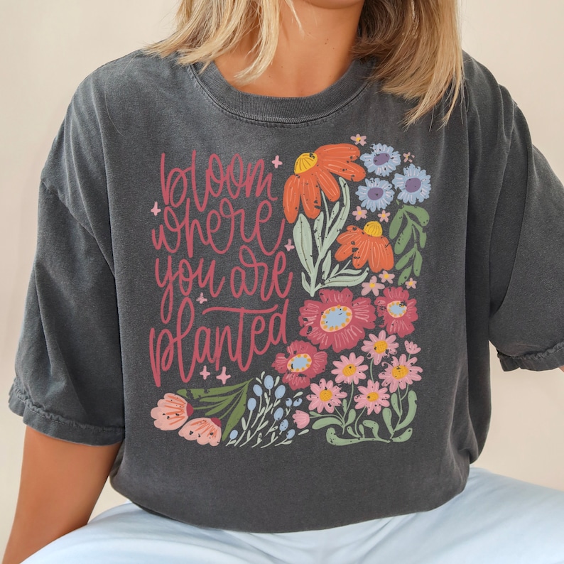 May include: A dark gray t-shirt with a floral design and the text "bloom where you are planted".