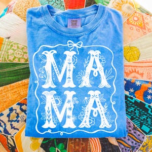 May include: A light blue t-shirt with a white floral design that says "MAMA" in a decorative font. The shirt is made by Comfort Colors.
