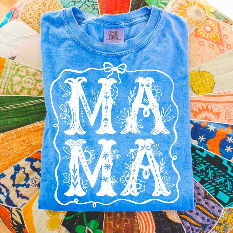 May include: A light blue t-shirt with a white floral design that says "MAMA" in a decorative font. The shirt is made by Comfort Colors.