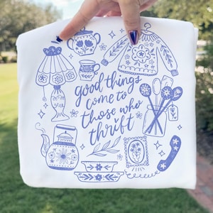 Op de afbeelding: Witte sweatshirt met een blauwe illustratie van diverse vintage items, waaronder een lamp, theepot en trui. De zin "good things come to those who thrift" is in blauwe letters geschreven.