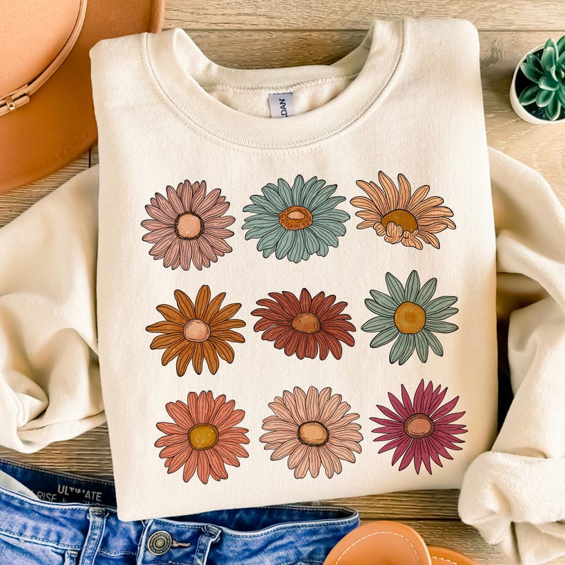 May include: A cream-colored sweatshirt with a pattern of nine colorful daisies. The daisies are arranged in three rows of three. The daisies are in shades of pink, orange, red, blue, and green.