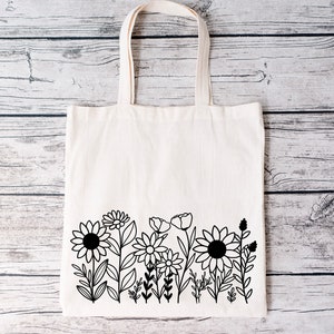 May include: A white canvas tote bag with black line drawings of flowers and leaves. The bag has a simple design and is perfect for carrying groceries or other items.
