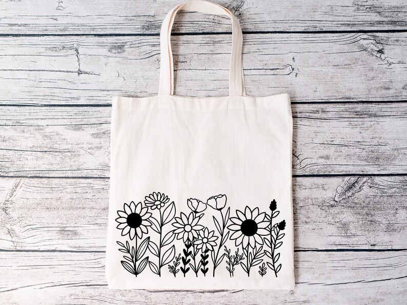 May include: A white canvas tote bag with black line drawings of flowers and leaves. The bag has a simple design and is perfect for carrying groceries or other items.