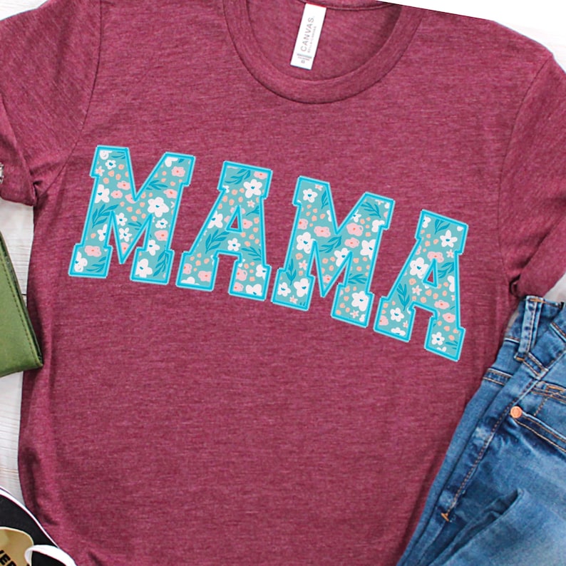 Op de afbeelding: Een maroon t-shirt met een bloemenpatroon dat het woord "MAMA" spelt in blauwe omrande letters.