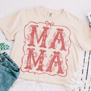 May include: A light beige t-shirt with a red floral design that says "MAMA" in a vintage style font.