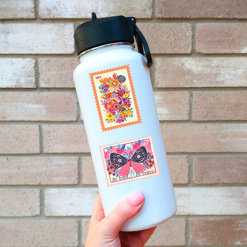 Pu&ograve; includere: Una bottiglia d'acqua in acciaio inossidabile bianca con due adesivi colorati. L'adesivo superiore presenta un mazzo di fiori con il testo "10&cent;". L'adesivo inferiore presenta una farfalla con il testo "Sii gentile. Sii coraggioso."