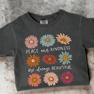 K&ouml;nnte beinhalten: Ein dunkelgraues T-Shirt mit einem grafischen Design aus bunten Blumen und dem Text "Peace and kindness are always beautiful".