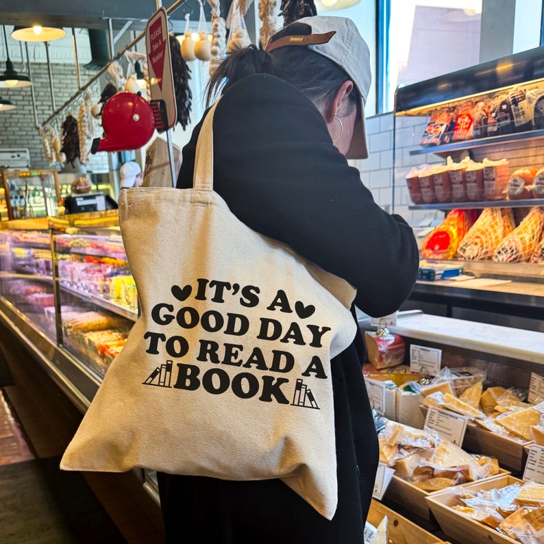 May include: A beige tote bag with the text "It's a good day to read a book" in black lettering. The bag has two small hearts on either side of the text and a graphic of stacked books below the text.