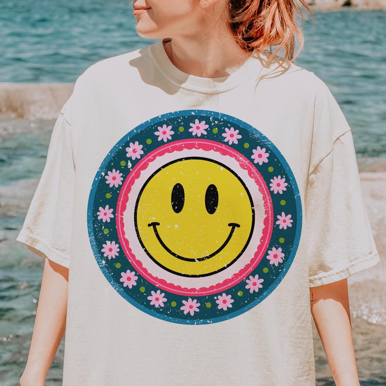 May include: A white t-shirt with a vintage-style smiley face graphic. The graphic features a yellow smiley face with a pink and blue border, surrounded by a ring of pink flowers on a blue background.