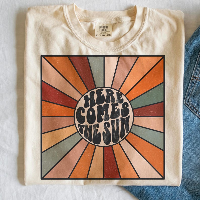May include: A beige t-shirt featuring a retro-style graphic of a sun with the text "Here Comes The Sun" in a circle. The sun graphic has a rainbow of colours radiating from it.