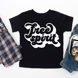 K&ouml;nnte beinhalten: Schwarzes T-Shirt mit dem Schriftzug "Free Spirit" in wei&szlig;er Retro-Schrift. Das Shirt wird mit einem karierten Button-Down-Hemd, einer blauen Jeans und schwarzen Leinenschuhen pr&auml;sentiert.