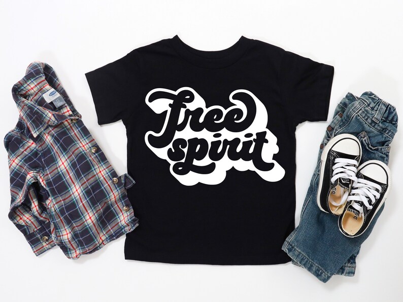 K&ouml;nnte beinhalten: Schwarzes T-Shirt mit dem Schriftzug "Free Spirit" in wei&szlig;er Retro-Schrift. Das Shirt wird mit einem karierten Button-Down-Hemd, einer blauen Jeans und schwarzen Leinenschuhen pr&auml;sentiert.