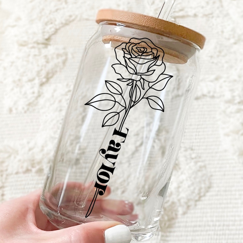 May include: A clear glass reusable drinking cup with a wooden lid and a black and white line drawing of a rose and the word "Taylor".