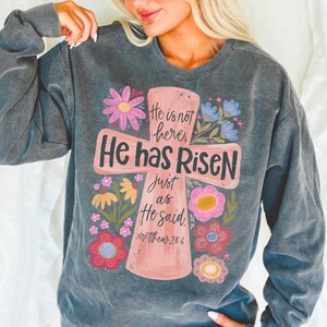 Puede incluir: Sudadera gris oscuro con un dise&ntilde;o de cruz rosa con las palabras "He is not here, He has Risen, just as He said" y el vers&iacute;culo b&iacute;blico Mateo 28:6. La cruz est&aacute; rodeada de flores coloridas en rosa, amarillo y azul.