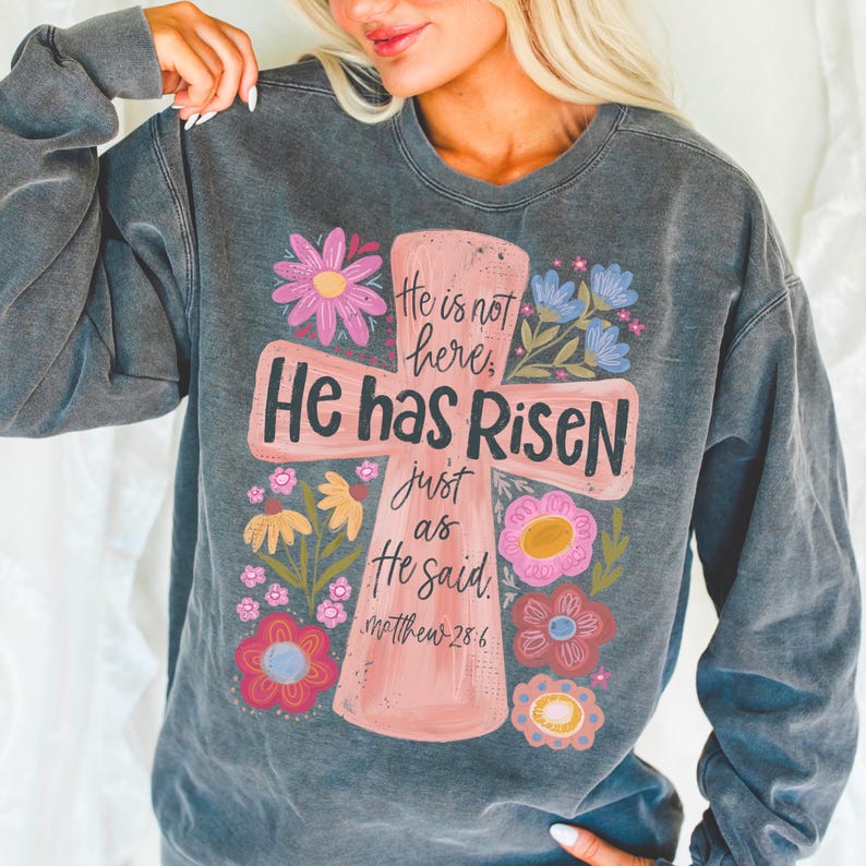 Puede incluir: Sudadera gris oscuro con un dise&ntilde;o de cruz rosa con las palabras "He is not here, He has Risen, just as He said" y el vers&iacute;culo b&iacute;blico Mateo 28:6. La cruz est&aacute; rodeada de flores coloridas en rosa, amarillo y azul.