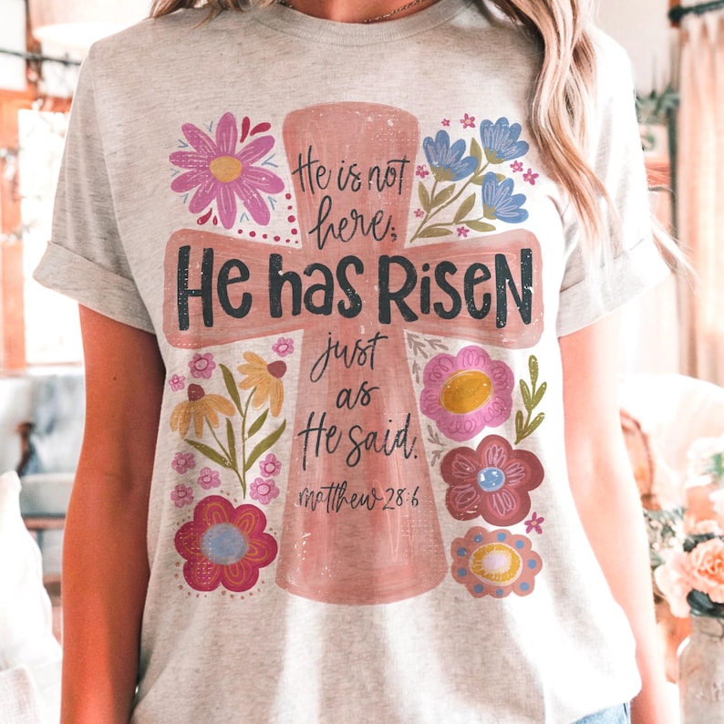 Puede incluir: Camiseta gris claro con un dise&ntilde;o de cruz rosa y las palabras "He is not here, He has Risen just as He said" y Mateo 28:6. La cruz est&aacute; rodeada de ilustraciones florales coloridas.
