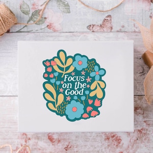 May include: A white card with a teal blue floral design and the text "Focus on the Good" in white.