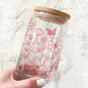 May include: A clear glass reusable water bottle with a pink floral design and a butterfly. The bottle has a bamboo lid and a straw.