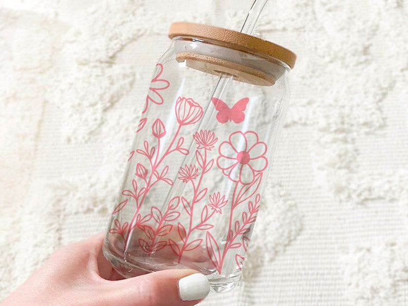May include: A clear glass reusable water bottle with a pink floral design and a butterfly. The bottle has a bamboo lid and a straw.