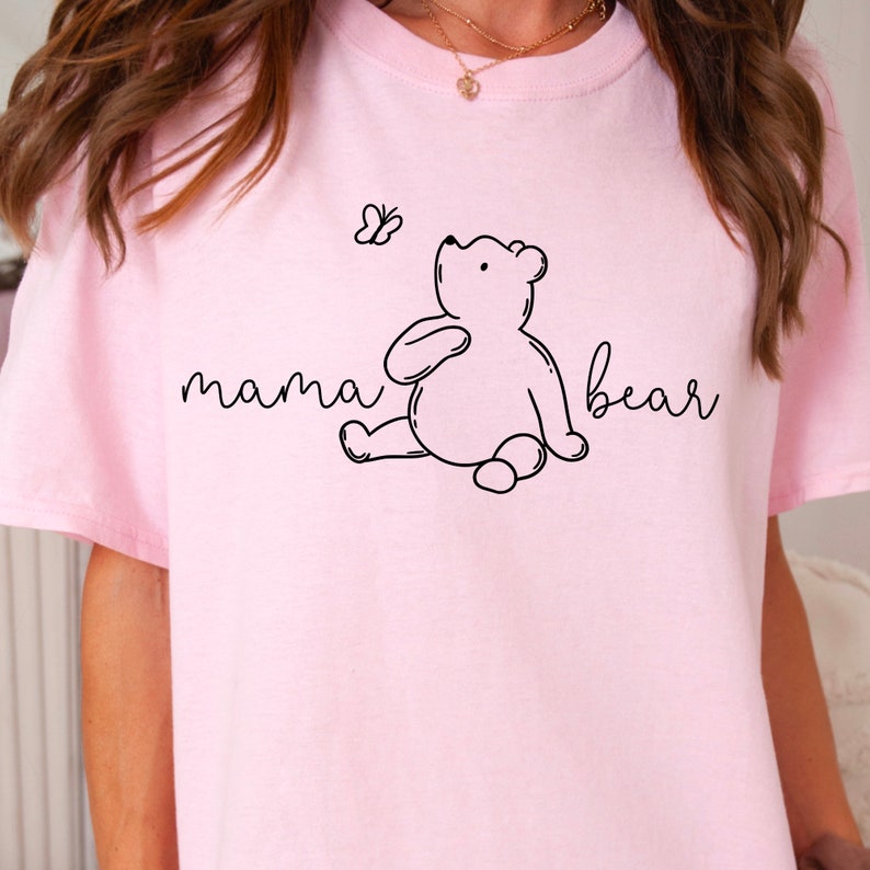 Puede incluir: Camiseta rosa con un contorno negro de una osa mam&aacute; y una mariposa. El texto "mama bear" est&aacute; escrito en cursiva.
