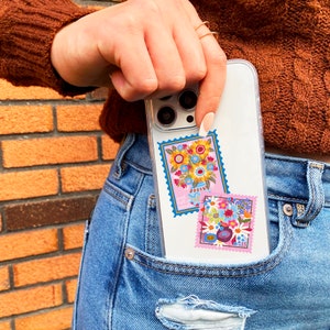 Pu&ograve; includere: Una custodia per telefono trasparente con un design floreale. La custodia ha due adesivi rosa e blu con disegni floreali. Il testo su un adesivo recita "Just full of happy vibes".