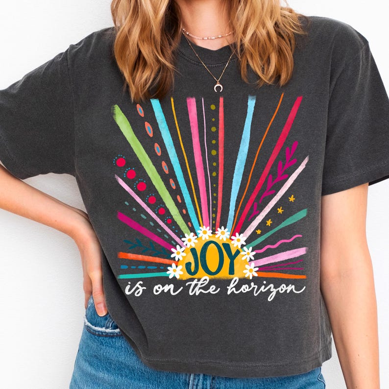 May include: A charcoal grey t-shirt with a colorful sunburst design and the text "JOY is on the horizon".