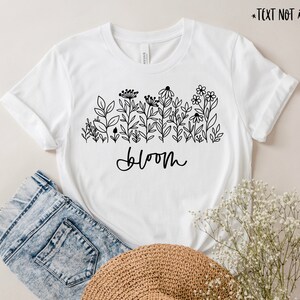 May include: White t-shirt with a black line drawing of flowers and the word "bloom" printed on the front.