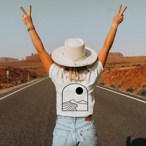 May include: A white t-shirt with a black desert landscape graphic and a sun. The person is wearing a white hat and blue jeans, standing on a road with arms raised in a peace sign. The background shows a desert landscape.