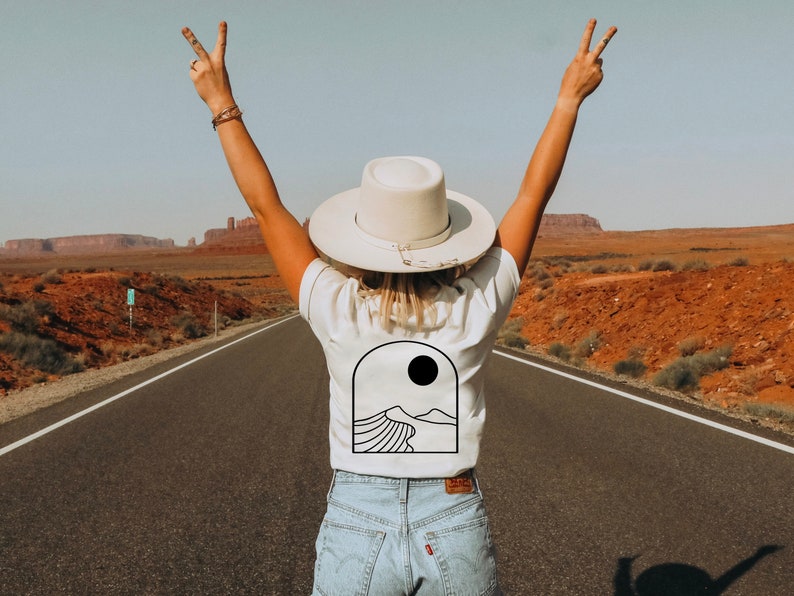 May include: A white t-shirt with a black desert landscape graphic and a sun. The person is wearing a white hat and blue jeans, standing on a road with arms raised in a peace sign. The background shows a desert landscape.