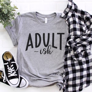 May include: A gray t-shirt with the text "Adult-ish" in black lettering. The shirt is laid flat on a white brick background with a black and white plaid shirt and black sneakers.