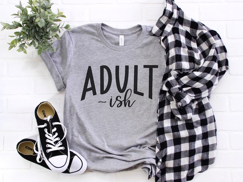 May include: A gray t-shirt with the text "Adult-ish" in black lettering. The shirt is laid flat on a white brick background with a black and white plaid shirt and black sneakers.
