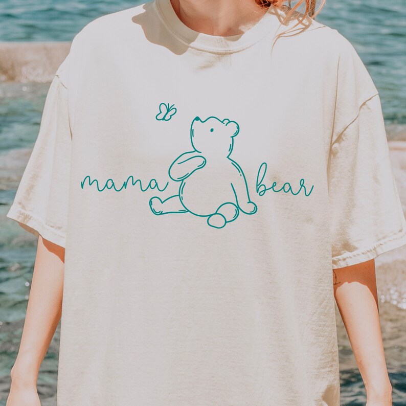 Puede incluir: Una camiseta blanca con un contorno azul verdoso de una osa madre y una mariposa. El texto "mama bear" est&aacute; escrito en cursiva azul verdoso.