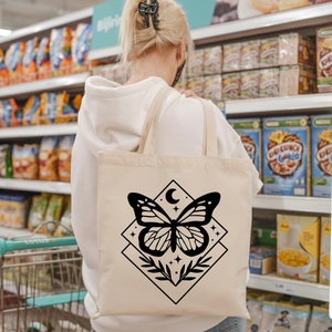 May include: A cream-coloured tote bag with a black butterfly design, a crescent moon, and stars. The butterfly is within a diamond shape with leaves. The bag is carried in a supermarket.
