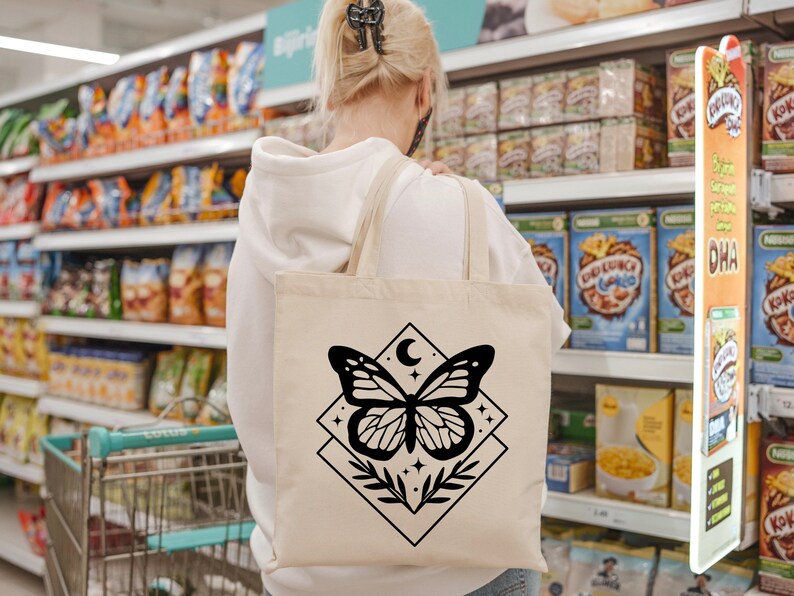 May include: A cream-coloured tote bag with a black butterfly design, a crescent moon, and stars. The butterfly is within a diamond shape with leaves. The bag is carried in a supermarket.