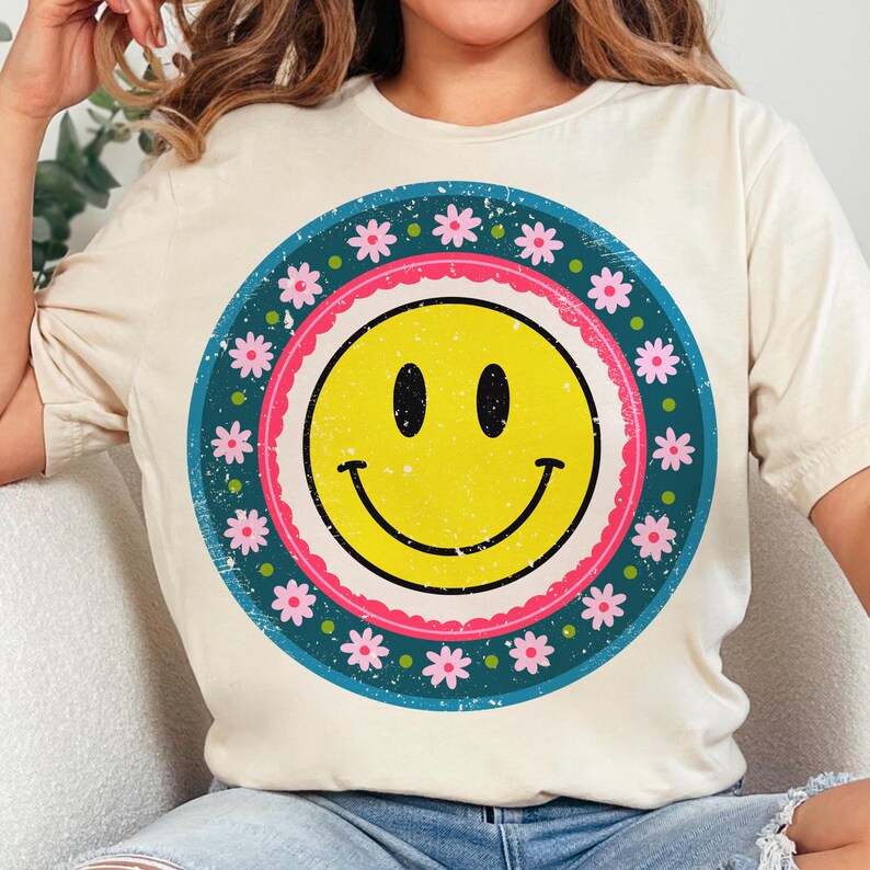 May include: A white t-shirt with a vintage-style graphic of a yellow smiley face surrounded by a blue circle with pink flowers.