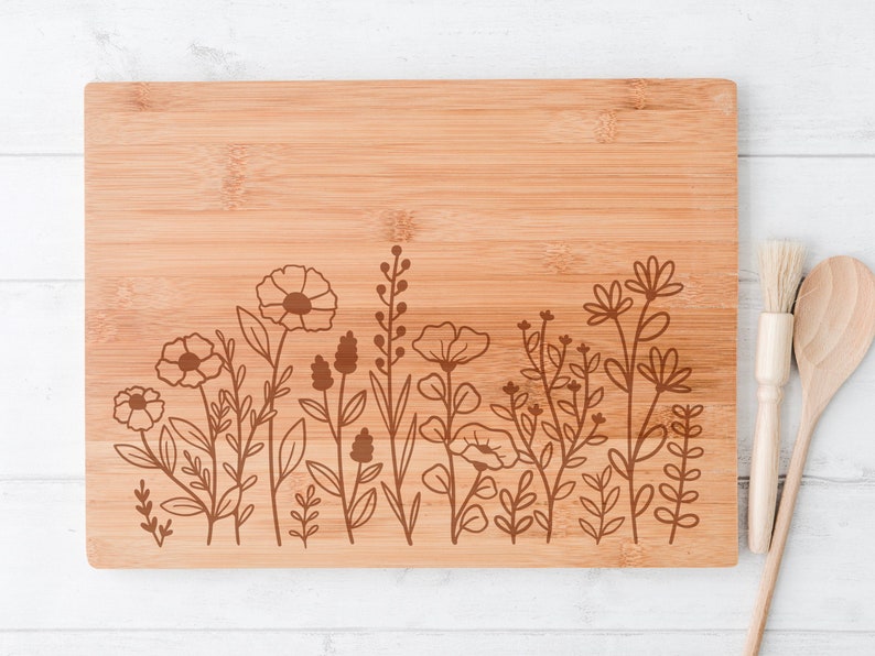 May include: A brown bamboo cutting board with a floral design engraved on the bottom edge. The design features a row of stylized flowers and leaves.