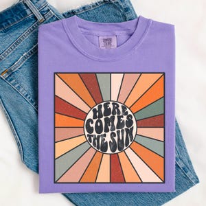 May include: Purple t-shirt featuring a retro sunburst design with the text "Here Comes The Sun" in a groovy font.