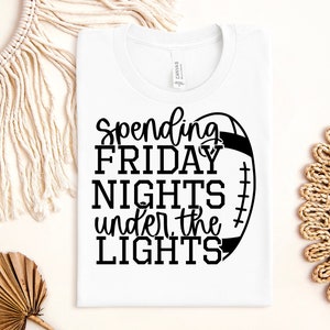 May include: White t-shirt with black text that reads "Spending Friday Nights Under the Lights" and a black and white football graphic.