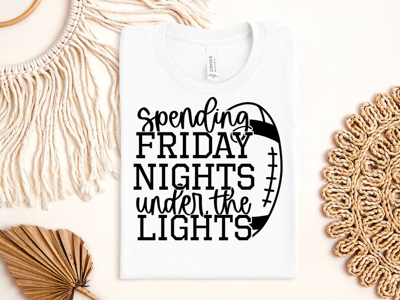 May include: White t-shirt with black text that reads "Spending Friday Nights Under the Lights" and a black and white football graphic.