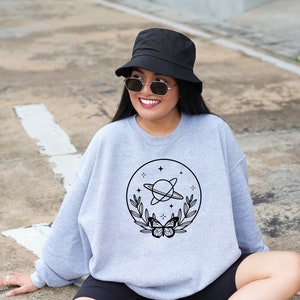 May include: A light grey sweatshirt with a black graphic of a planet with rings, stars, leaves, and a butterfly. The person in the image is wearing a black bucket hat and sunglasses.
