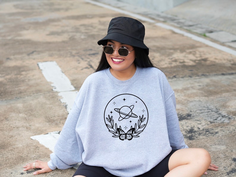May include: A light grey sweatshirt with a black graphic of a planet with rings, stars, leaves, and a butterfly. The person in the image is wearing a black bucket hat and sunglasses.