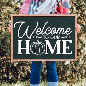 May include: A person holding a black chalkboard sign with white text that says "Welcome to our Home" with a white pumpkin graphic.