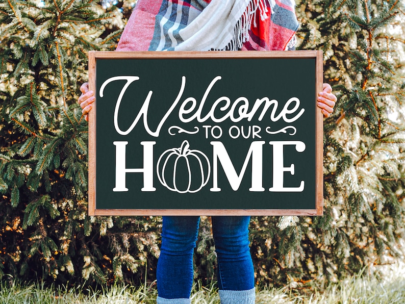 May include: A person holding a black chalkboard sign with white text that says "Welcome to our Home" with a white pumpkin graphic.