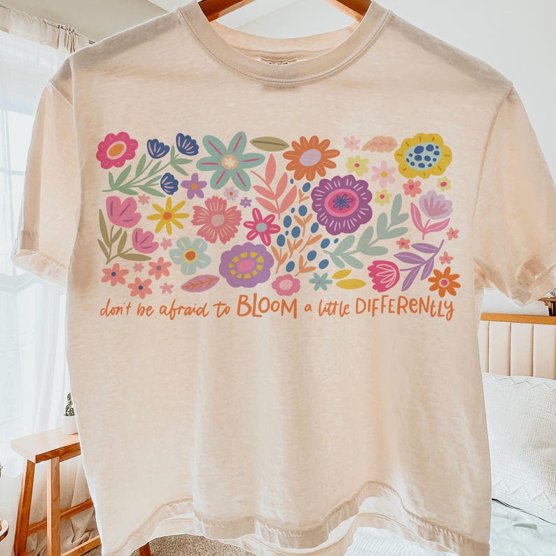 May include: A light pink cropped t-shirt with a colorful floral design and the text "don't be afraid to BLOOM a little DIFFERENTLY".